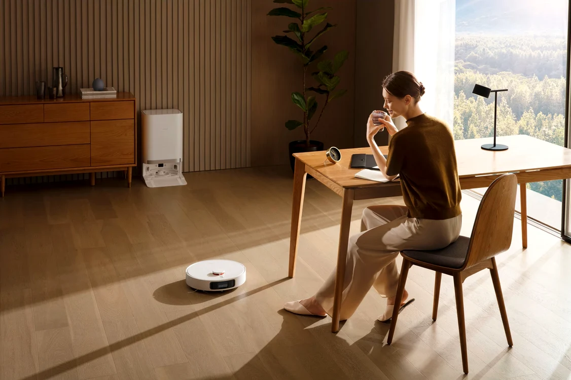 Femeie la cafea în timp ce aspiratorul robot curăță livingul