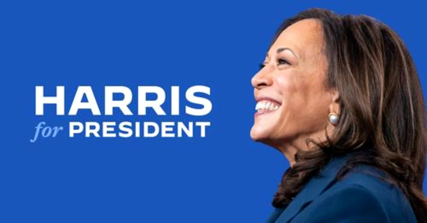 Kamala Harris zâmbind lângă un afiș cu numele său