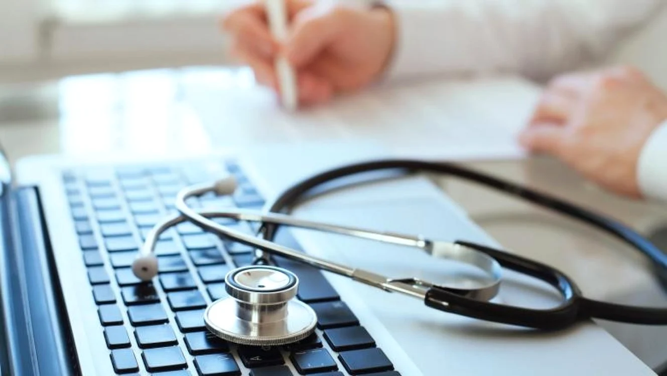 Medic care scrie o rețetă în cabinetul medical, cu stetoscopul lăsat pe tastatura laptopului