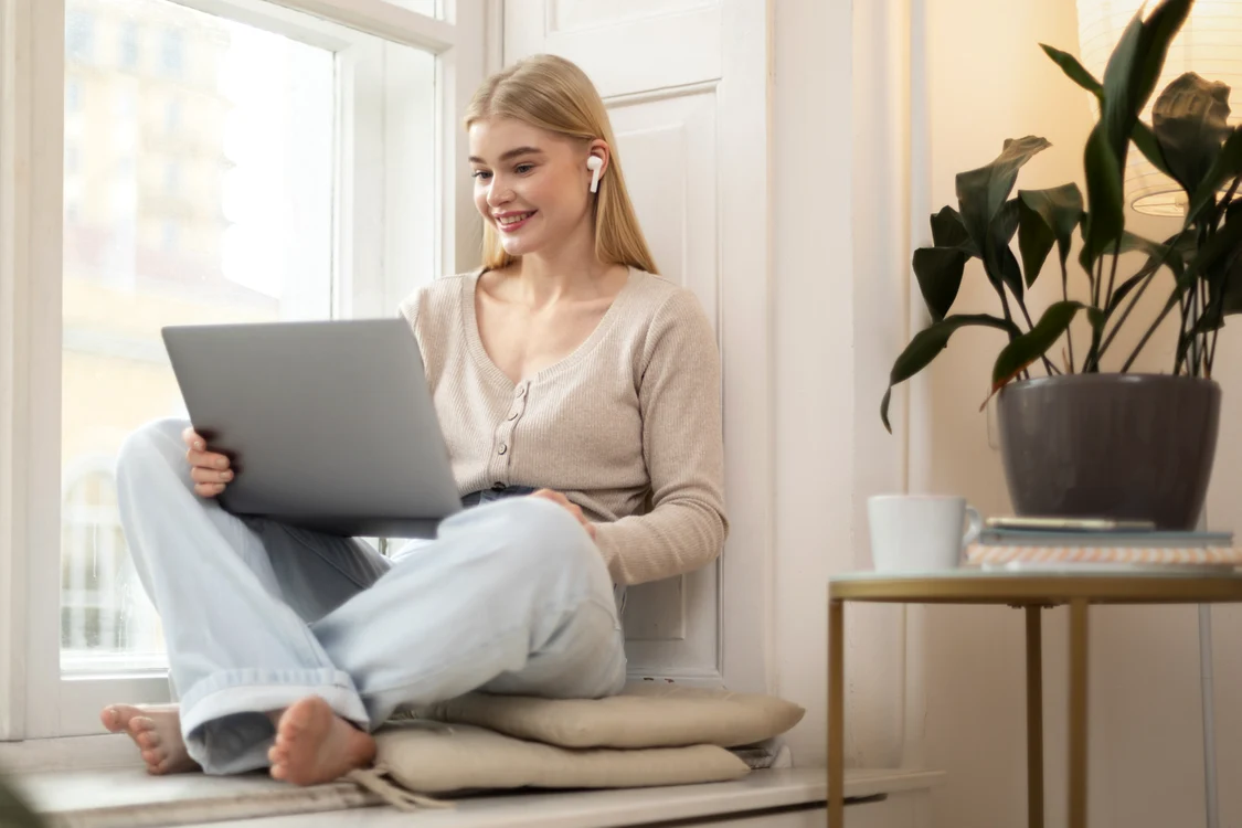 O tânără lucrează la laptop stând pe pervazul foarte lat al ferestrei