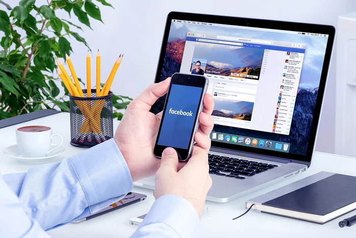 Un laptop și un telefon in mana cu pagina de Facebook