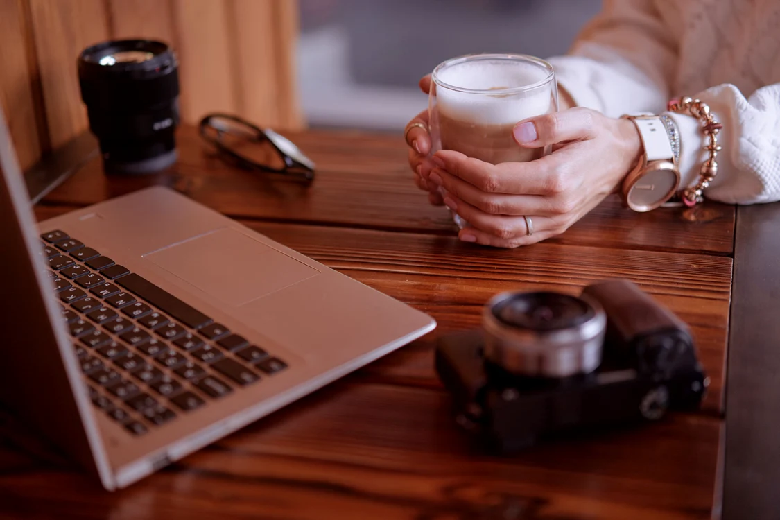 O femeie ține între palme un pahar cu cafea cu lapte în timp ce citește la un laptop pus pe birou