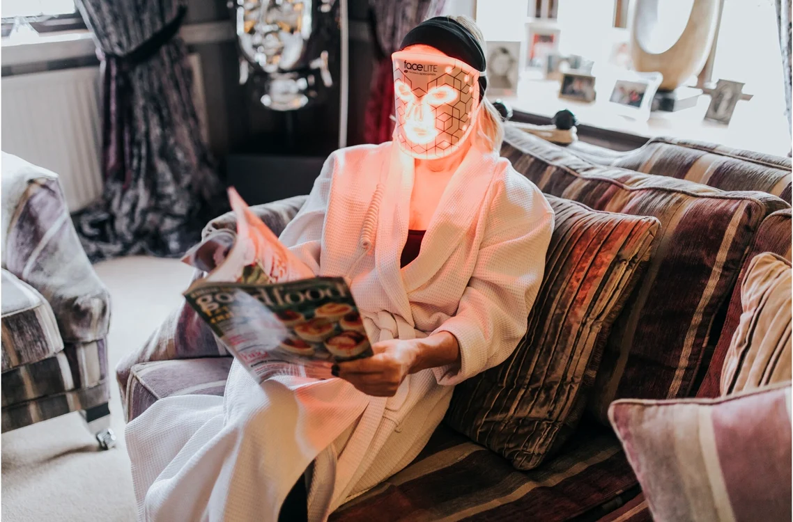 Femeie relaxată în fotoliu, citind o revistă în timp ce folosește o mască facială cu LED acasă
