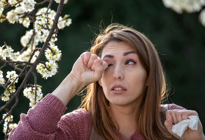Femeie care se scarpină la ochi din cauza alergiilor sezoniere, lângă un copac înflorit Adevărul