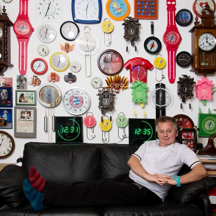 Werner Stechbarth fotografiat pe o canapea, în fața unui perete cu colecția lui de ceasuri