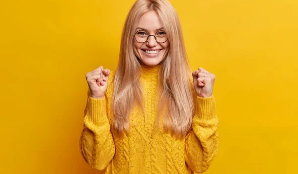 happy joyful blonde woman clenches fists celebrates good result smiles broadly being high spirit makes triumph gesture wears spectacles winter sweater jpg