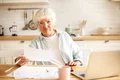 elderly housewife with gray hair sitting kitchen with open laptop papers table having emotional frustrated facial expression shocked with debt amount while paying domestic bills online jpg