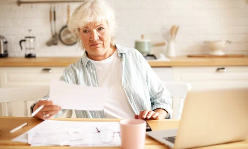 elderly housewife with gray hair sitting kitchen with open laptop papers table having emotional frustrated facial expression shocked with debt amount while paying domestic bills online jpg