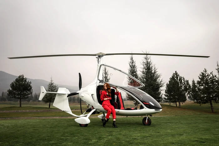 Girocopter la sol, pe marginea căruia stă o femeie îmbrăcată în roșu