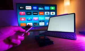 o persoana opereaza telecomanda unui mediaplayer langa un laptop si un televizor in fundal jpg