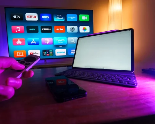 o persoana opereaza telecomanda unui mediaplayer langa un laptop si un televizor in fundal jpg