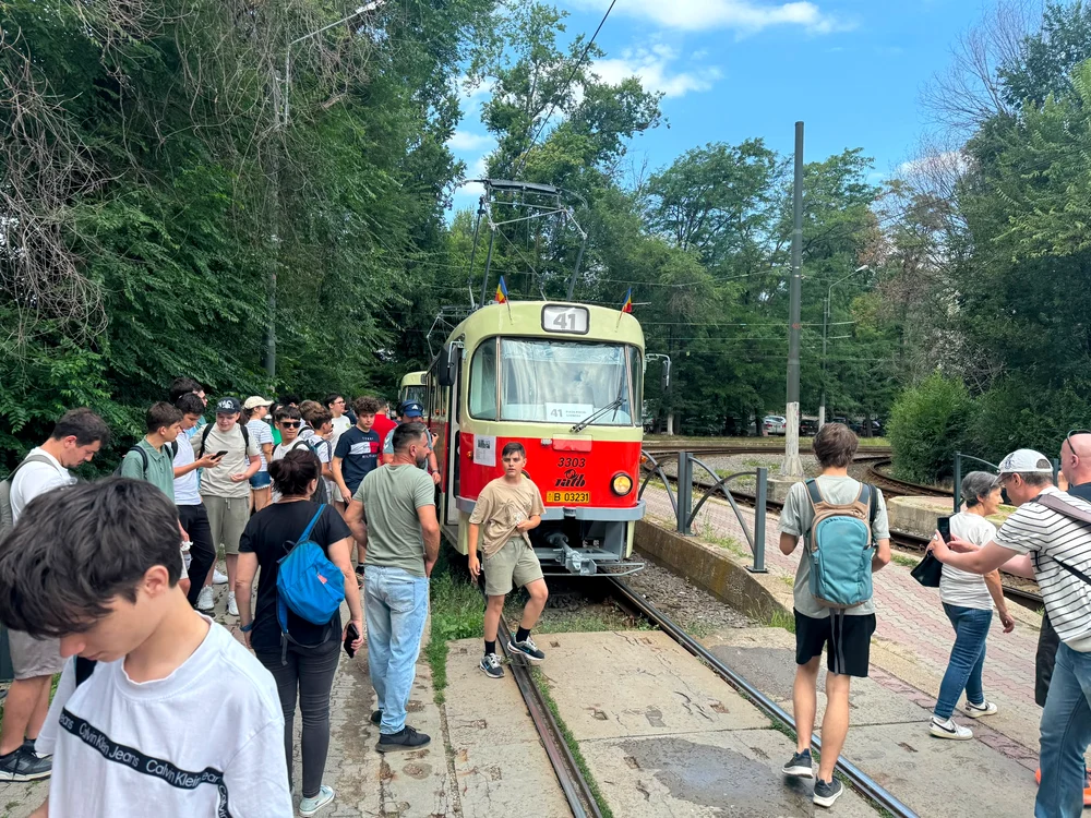 Zeci de oameni, printre care mulți tineri, mergând pe lângă șinele de tramvai spre un vagon Tatra