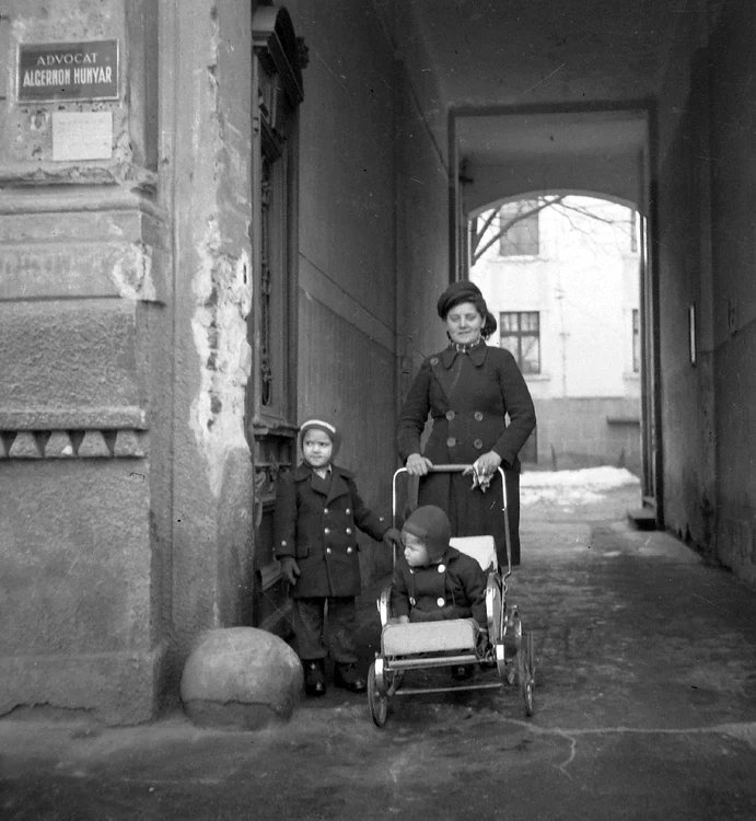 Femeie cu doi copii, unul în cărucior, într-o curte urbană veche.