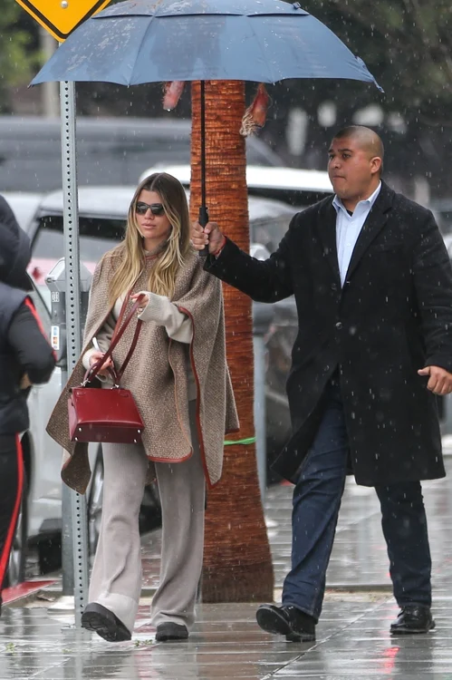 Sofia Richie, îmbrăcată în bej mergând prin ploaie. Un asistent personal îi ține umbrela