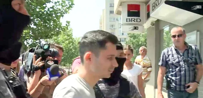 
    Tânărul de 22 de ani  a fost adus  încătușat la  sediul bănciiCaptura: Digi 24  