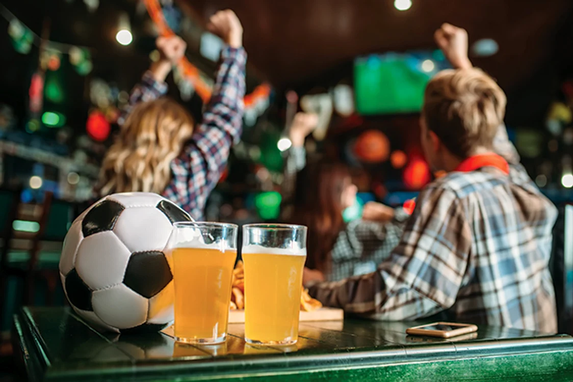 Două pahare pline cu bere, o minge de fotbal și suporterii care sărbătoresc