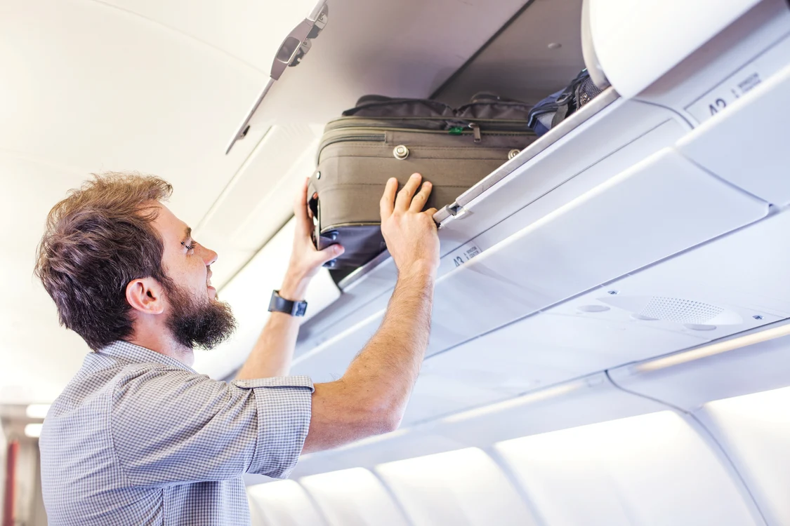 Un bărbat care-și așază bagajul de mână în compartimentul de deasupra locului său din avion Shutters