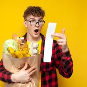 Student ținând în brațe o pungă cu diverse cumprături, fiind șocat de bonul de cumpărături shutterstock jpg