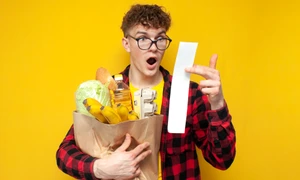 Student ținând în brațe o pungă cu diverse cumprături, fiind șocat de bonul de cumpărături  shutterstock jpg