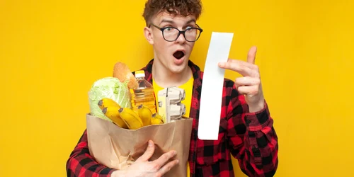 Student ținând în brațe o pungă cu diverse cumprături, fiind șocat de bonul de cumpărături  shutterstock jpg