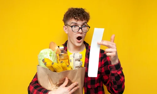 Student ținând în brațe o pungă cu diverse cumprături, fiind șocat de bonul de cumpărături  shutterstock jpg
