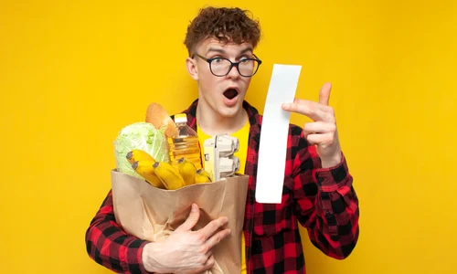 Student ținând în brațe o pungă cu diverse cumprături, fiind șocat de bonul de cumpărături  shutterstock jpg