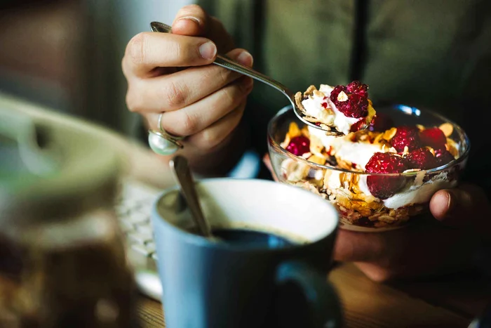 O doamnă care mănâncă cereale cu iaurt și fructe și are lângă o cană cu cafea la micul dejun