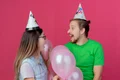 surprised young couple wearing party hat looks each other standing with helium balloons isolated pink wall jpg
