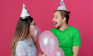 surprised young couple wearing party hat looks each other standing with helium balloons isolated pink wall jpg
