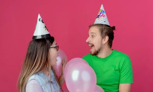 surprised young couple wearing party hat looks each other standing with helium balloons isolated pink wall jpg