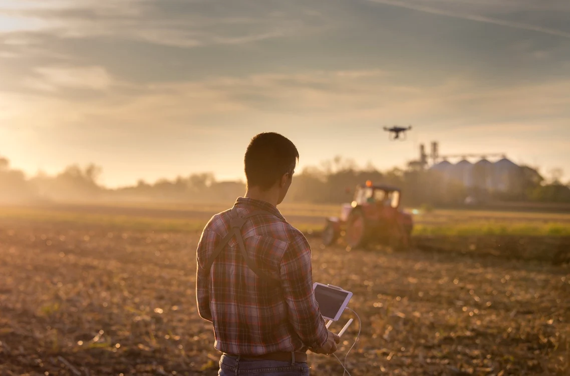 Un tânăr fermier ghidează o dronă deasupra unui tractor care ară pe câmp