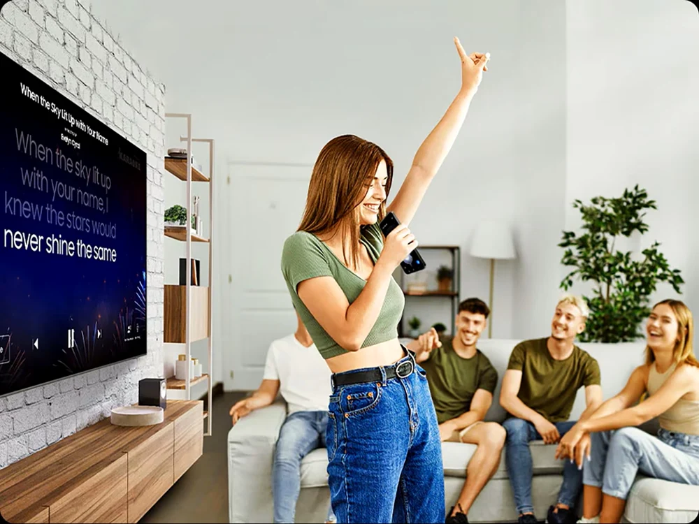 Grup de prieteni face karaoke în sufragerie, cu versurile afișate pe un televizor QLED