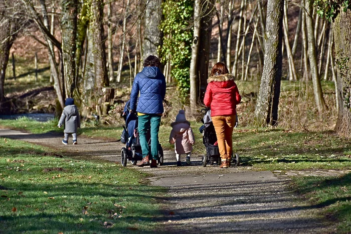 Două mămici își plimbă bebelușii în parc, cu căruciorul, în timp ce alți doi copii merg pe lângă ele