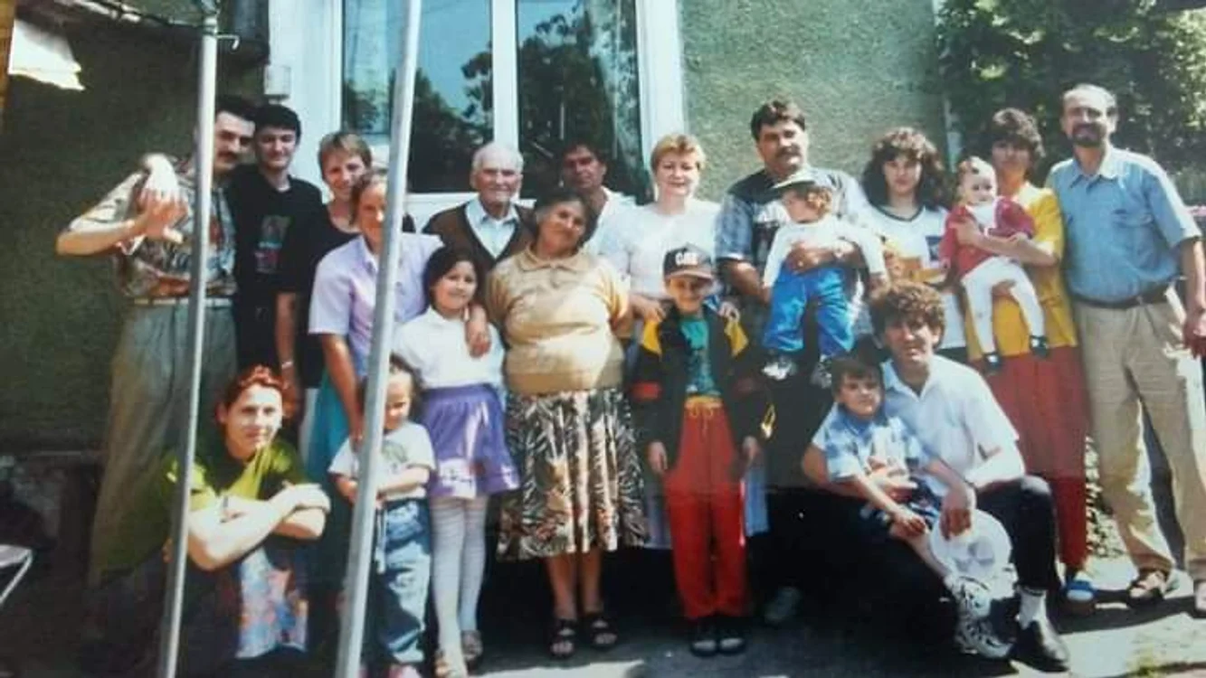 Smiley în copilărie, îmbrăcat în pantaloni roșii, tricou, geacă sport, cu papuci și șapcă, alături de bunica lui și de familie