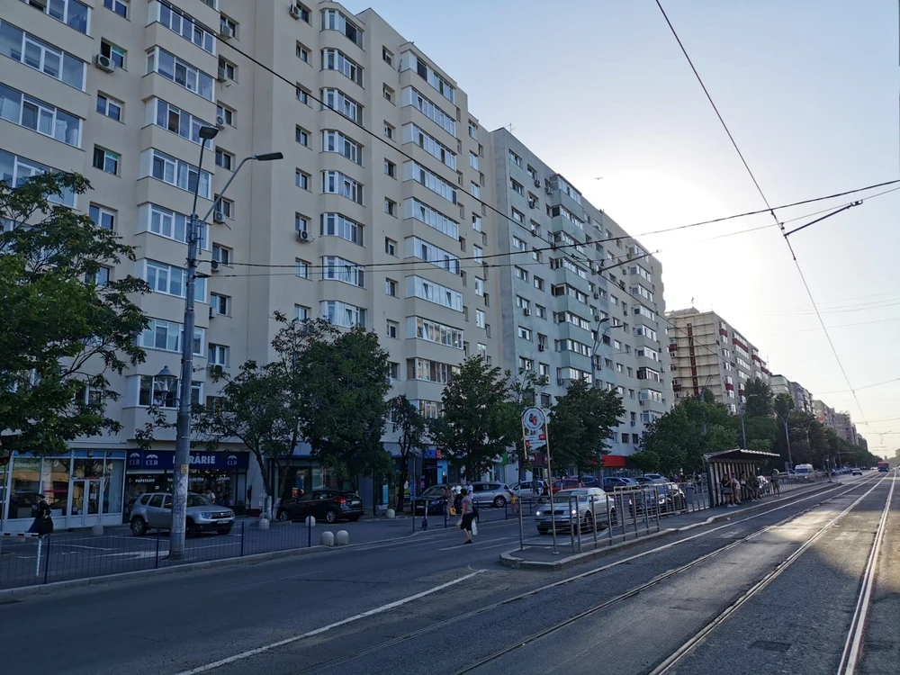 Bloc de locuințe cu multe etaje, pe o stradă cu linie de tramvai, copaci și mașini parcate, fotograf
