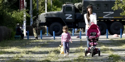 20 mai 2022 - o femeie şi un copil se plimbă într-un parc în timp ce un soldat rus patrulează pe străzile din  Skadovsk regiunea Herson FOTO AFP 