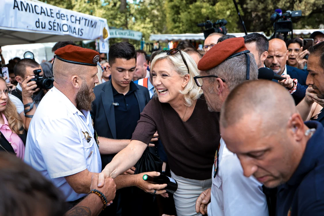 Marine Le Pen dă mâna cu un bărbat purtând beretă militară, înconjurată de susținători