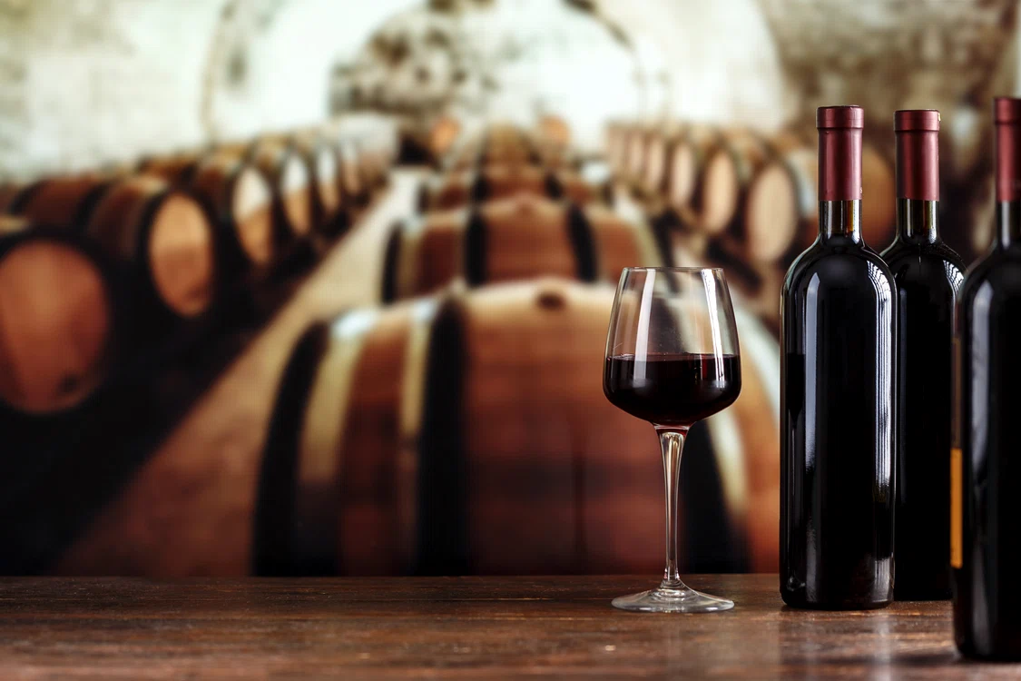 Cramă cu foarte multe butoaie cu vine și câteva sticle de vin roșu pe o masă, lângă un pahar