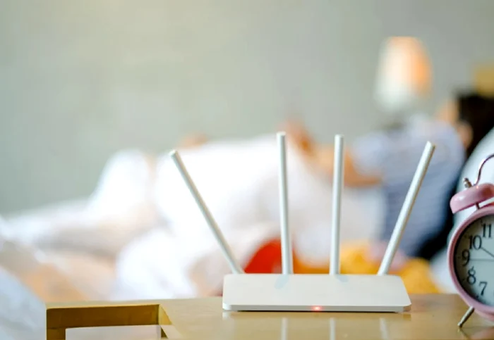 Un router pe un birou, alături de un ceas, și o persoană care doarme în pat, în fundal Shutterstock