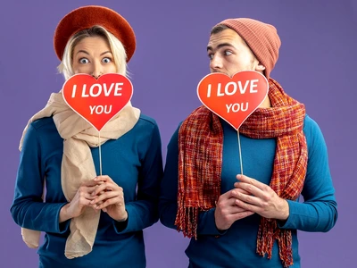 young couple wearing hat with scarf valentine s day covered face with red hearts stick with i love you text pleased guy looking girl isolated blue background jpg