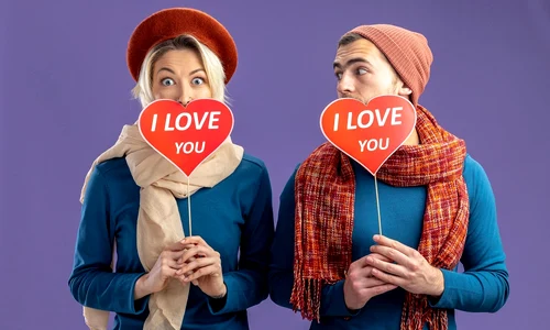 young couple wearing hat with scarf valentine s day covered face with red hearts stick with i love you text pleased guy looking girl isolated blue background jpg