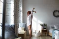 full lenght shot attractive young redhead female long striped shirt standing barefooted her toes bedroom stretching body raising arms facing big window enjoying warm spring sunlight jpg