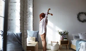 full lenght shot attractive young redhead female long striped shirt standing barefooted her toes bedroom stretching body raising arms facing big window enjoying warm spring sunlight jpg