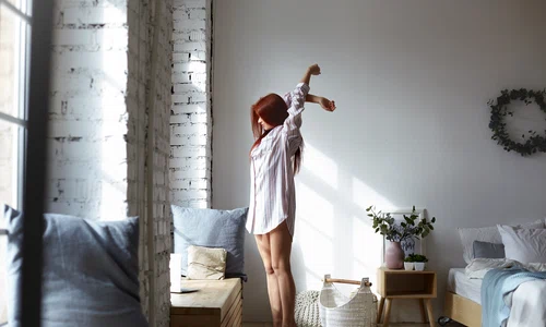 full lenght shot attractive young redhead female long striped shirt standing barefooted her toes bedroom stretching body raising arms facing big window enjoying warm spring sunlight jpg
