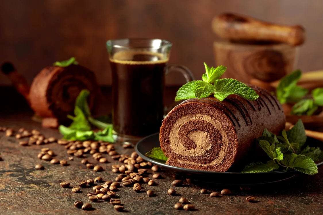 O cană de cafea și o ruladă cu cremă pe o masă în care sunt împrăștiate boabe de cafea Adevărul