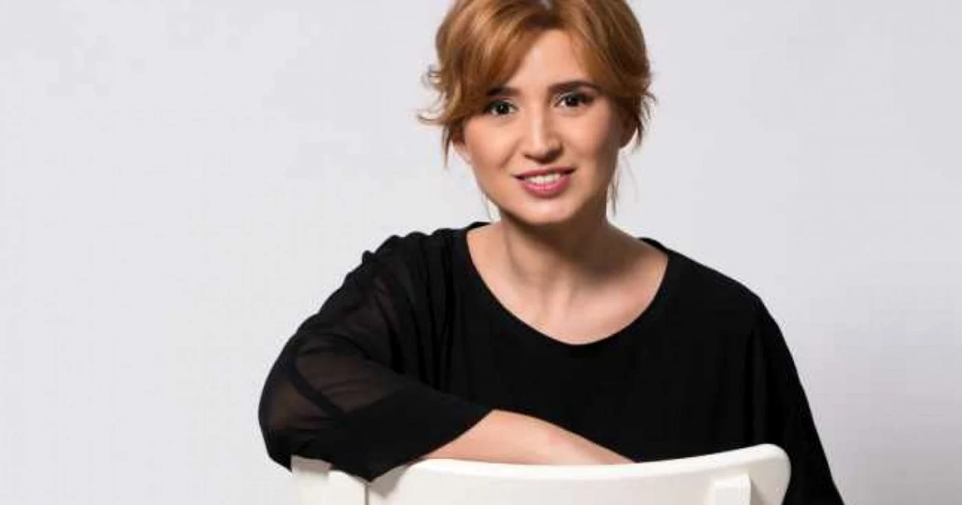 Sanda Nicola, fosta jurnalistă, îmbrăcată într-o bluză neagră stând pe un scaun alb