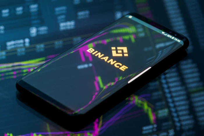 Telefon pe care este deschisă aplicația Binance, deasupra unui ecran cu grafice bursiere