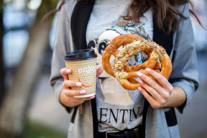 Studentă ținând în mâini un pahar de cafea și doi covrigi